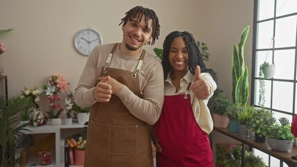 A man and woman florists in aprons standing in a flower shop, giving thumbs up, surrounded by plants and flowers with a cheerful demeanor.