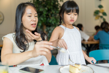 A twenty-something Chinese Malaysian woman and a six-year-old girl enjoying cake together at a stylish cafe in Selangor, Malaysia, on a sunny early summer afternoon.