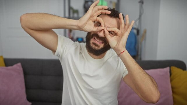 Cheeky middle-aged man at home, wearing white tee, goofing around by peeping through ok-sign fingers! eyes looking, tongue sticking out; a fun, madcap vision!