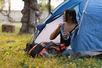 Thirsty young woman is sitting in a blue tent at a campsite, drinking from a bottle of water, relaxing and enjoying traveling to a natural outdoor environment.