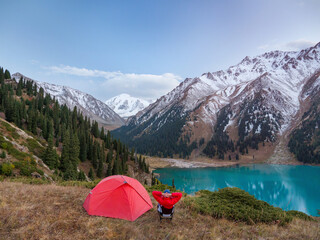 A male hiker sitting on a folding chair near a tent in a high mountainous area. He enjoys a magnificent panoramic view of snow-capped peaks and a turquoise mountain lake