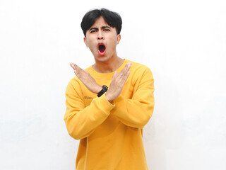 Portrait of an Asian young adult man wearing a yellow sweater, showing an angry expression and making a stop gesture with his hand, isolated on a white background.