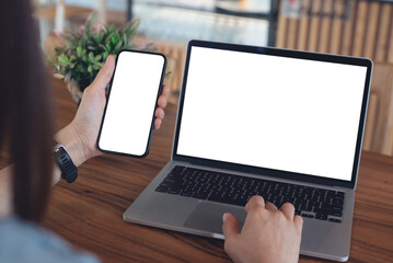 Mockup blank white screen cell phone and laptop for web template or mobile app design. Woman hand using mobile phone during working on laptop computer on wooden table at coffee shop or home office