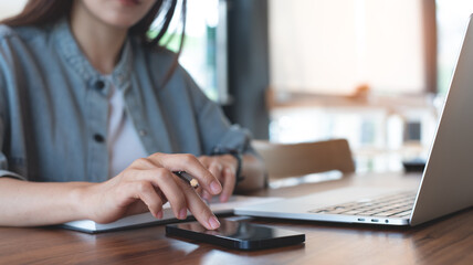 Young asian business woman using mobile phone on table during working on laptop computer at coffee shop. Female student studying online class, e-learning, using smartphone, remote working