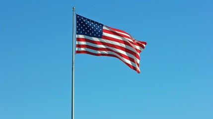 Fototapeta premium An American flag is waving prominently against a clear blue sky. The flag is mounted on a flagpole with a gold finial at the top. The flag's red, white, and blue colors are vibrant and the stars and s