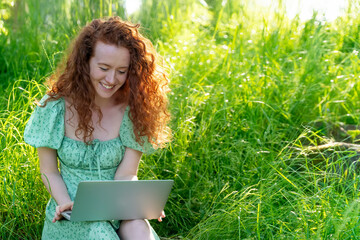 Smiling Woman With Red Hair Using Laptop in  Meadow on Sunny Day