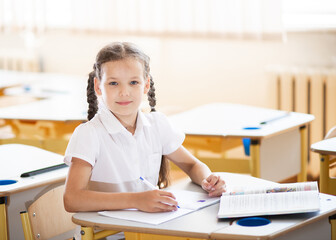 Back to school, girl sitting at a desk in the classroom. A schoolgirl sits at her desk and laughs joyfully; the child has school supplies on the table. The girl is dressed in a school uniform.
