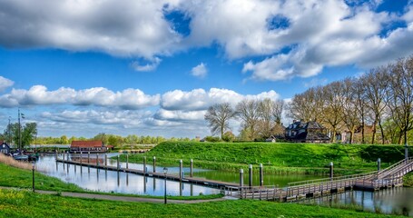 a peaceful river with a wooden dock, green grass, and houses under a blue sky with fluffy clouds.
