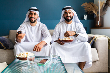Arab men wearing traditional emirate clothing  eating pop-corn and watching TV at home - Middle...