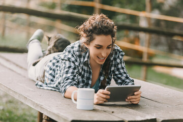 Young woman with curly hair in a braid, dressed in a checkered shirt and light pants, smiles while lying on a bench with a smartphone. A cat rests on her back, adding to the relaxed atmosphere