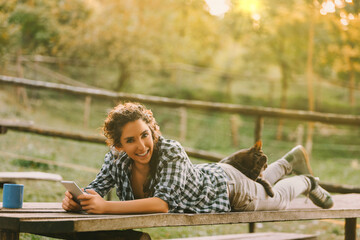 Naklejka premium Woman with braided curly hair, wearing a checkered shirt, holds a smartphone and smiles while lying on a bench. A cat resting on her back adds to the relaxed, outdoor vibe