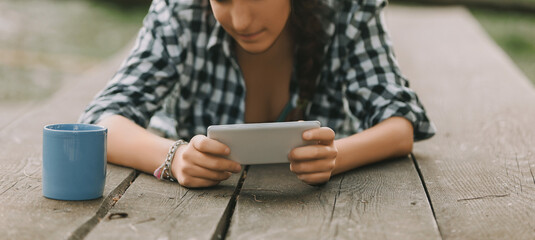 woman with curly braided hair and a checkered shirt lies on her stomach, engrossed in a smartphone....