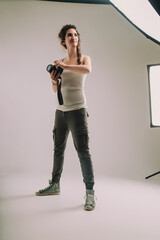 Young woman with curly braided hair, dressed in a sleeveless top and cargo pants, holds a camera in a studio. Her confident stance and expression reflect her passion and pride in photography