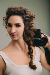 Young woman with curly hair in a braid, wearing a sleeveless top, carries a camera on her shoulder....