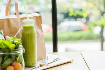 A glass bottle filled with a vibrant green smoothie sits beside a salad, both waiting to be enjoyed. A brown paper bag rests beside them, suggesting a recent takeout order.
