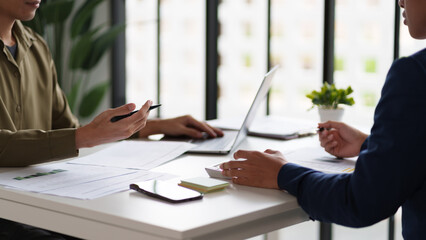  professional setting featuring two individuals working at a table. They are engaged in reviewing documents and using a calculator. Laptops are open on the table.