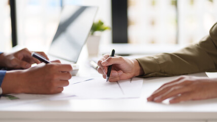 Close-up of two individuals discussing and pointing at a document on a desk. The hands are holding pens, indicating a review or collaboration on the content of the document