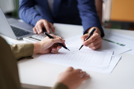 Close-up of two individuals discussing and pointing at a document on a desk. The hands are holding pens, indicating a review or collaboration on the content of the document