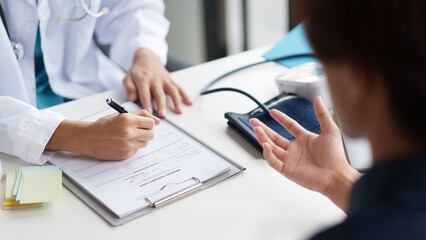 A doctor and patient during a consultation. The doctor is writing on a clipboard while the patient is explaining their symptoms. Medical bottles and a blood pressure monitor are visible on the table