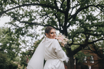 Wedding photography, portrait of a beautiful young happy smiling bride in white lingerie with a bouquet of flowers in a park outdoors against a background of green vegetation, nature.