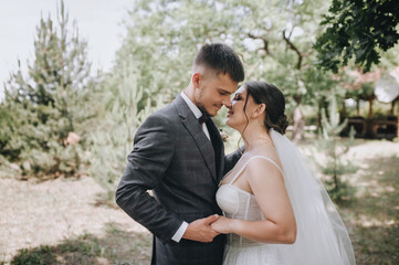 Fototapeta premium Handsome young groom in a suit and a bride in a white dress are hugging outdoors in the park. Photography, portrait of stylish newlyweds.