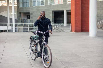 Fototapeta premium African businessman talking on cellphone while walking with electric bike in financial district