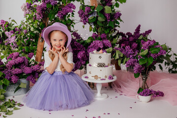A little blonde girl in a white blouse and a lilac skirt playfully holds a lilac hat in her hands. There is a white birthday cake nearby.