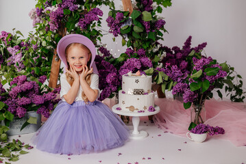 A little blonde girl in a white blouse and a lilac skirt playfully holds a lilac hat in her hands. There is a white birthday cake nearby.