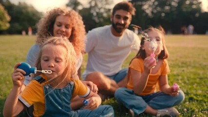 A happy family of parents and their two daughters are having a joyful time in the park, making bubbles together on a sunny day, and smiling with love
