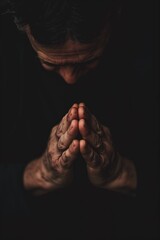 Person in contemplative pose with hands clasped together against dark background. Close-up shot of individual praying or meditating. Focus on subject actions and emotions.