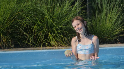 A teenage girl in a pool holding a ball and laughing, enjoying a sunny day.