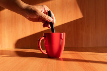 The hand of the Latin man. Hand moving liquid with a spoon in a red cup on a wooden background with daylight. Red cup. Corporate color. ceramic mug
