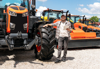Caucasian adult man with digital tablet in hand stands next to construction or agricultural tractor at a machinery dealer. Ag equipment dealer or supplier. © Barillo Images
