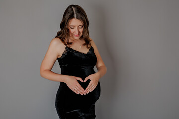 Fototapeta premium Pregnant girl in black dress on grey background stroking belly