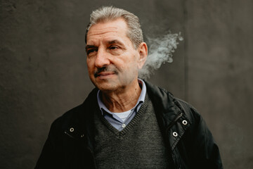 Older Man With Mustache Exhaling Smoke In Front Of Gray Wall