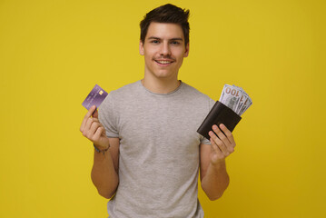 A young man smiles cheerfully as he displays a credit card and cash on a bright yellow background