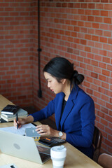 A businesswoman in a blue suit working at a desk with a laptop, holding a pen and writing on a document. A small model house and coffee cup are placed on the desk