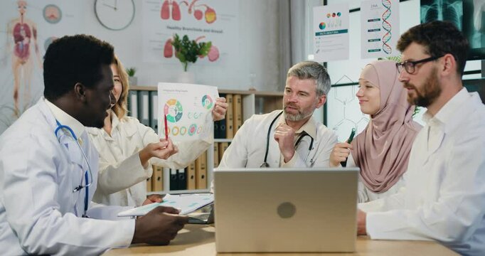 Multicultural medical colleagues man and woman doctors discussing results of researches with partners via video chat on laptop during joint meeting in clinic