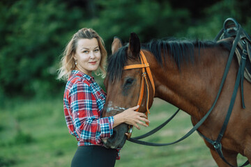 Obraz premium Happy blonde with horse in forest. Woman and a horse walking through the field during the day. Dressed in a plaid shirt and black leggings.