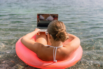 A woman is sitting on a red inflatable raft with a laptop on her lap. She is wearing a white bikini and sunglasses. Concept of relaxation and leisure.