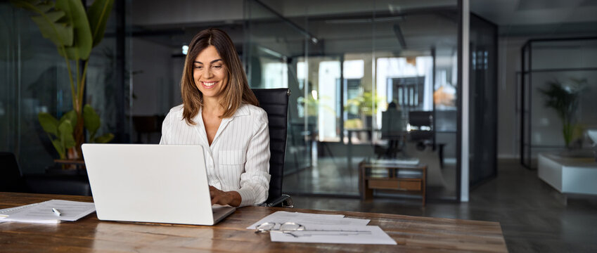 Middle age European business woman CEO using laptop application for work at table workspace in office. Smiling Latin Hispanic mature adult professional businesswoman using pc digital computer. Banner - Powered by Adobe