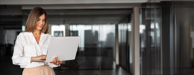 Banner of hispanic mature adult professional business woman looking at online trade app. European businesswoman CEO holding pc laptop computer using fintech application standing in office. Copy space
