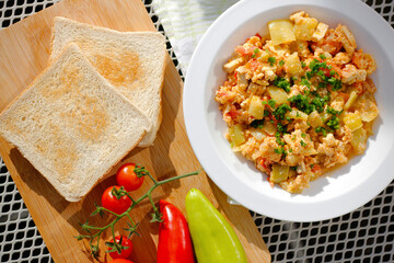 Vegetable dish called lecho containing peppers, tomatoes, onion, eggs and tofu served on white plate with chopped chives and toasted bread. Delicious lecho and its ingredients on garden table.