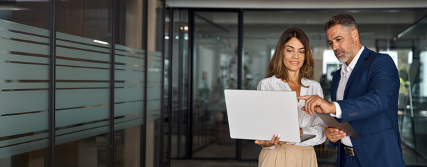 Mature mentor man and European leader woman discussing project on laptop in office. Two diverse...