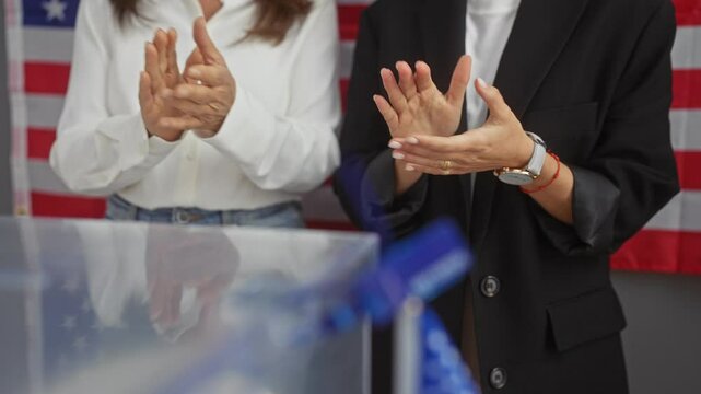 Two women wearing business attire applauding in an indoor setting with an american flag in the background, suggesting an electoral or political event.