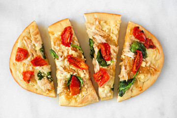 Flat bread pizza with chicken, parmesan cheese, sundried tomatoes, spinach and caramelized onions. Top down view on a white marble background.