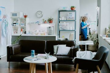 Black leather sofa and dark velvet armchair with small wooden coffee table between them, nurse preparing for work in blurred background