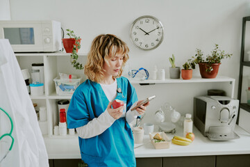 Young pretty Caucasian nurse eating red apple while communicating via smartphone during her break...