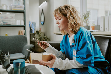 Pretty blonde Caucasian nurse checking her subscription and holding cardboard container with salad while having break in on call room