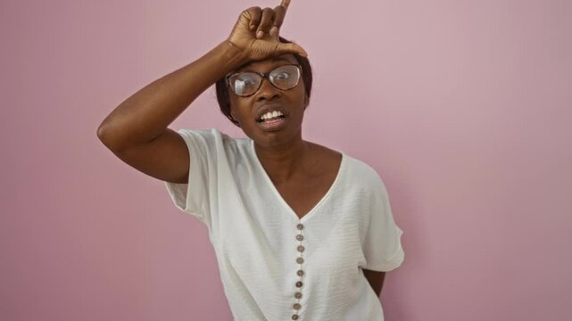 Young african american woman in glasses standing with loser finger gesture on forehead mocking people over isolated pink background insulting and teasing with a smile
